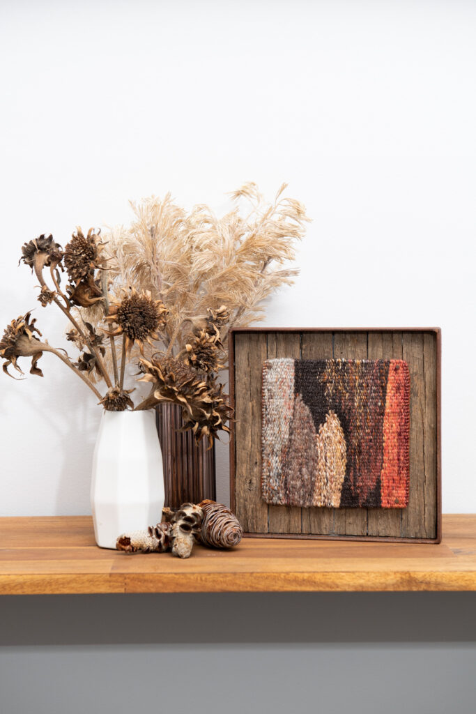A small earthen coloured tapestry sits beside dried flower arrangements on a shelf