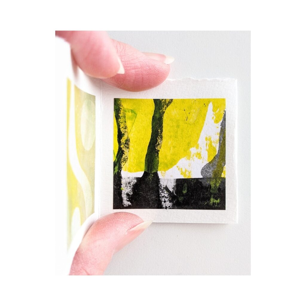 A tiny yellow, white and black painting is held between a  woman's fingers and thumb. 