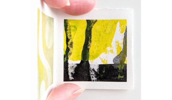 A tiny yellow, white and black painting is held between a woman's fingers and thumb.