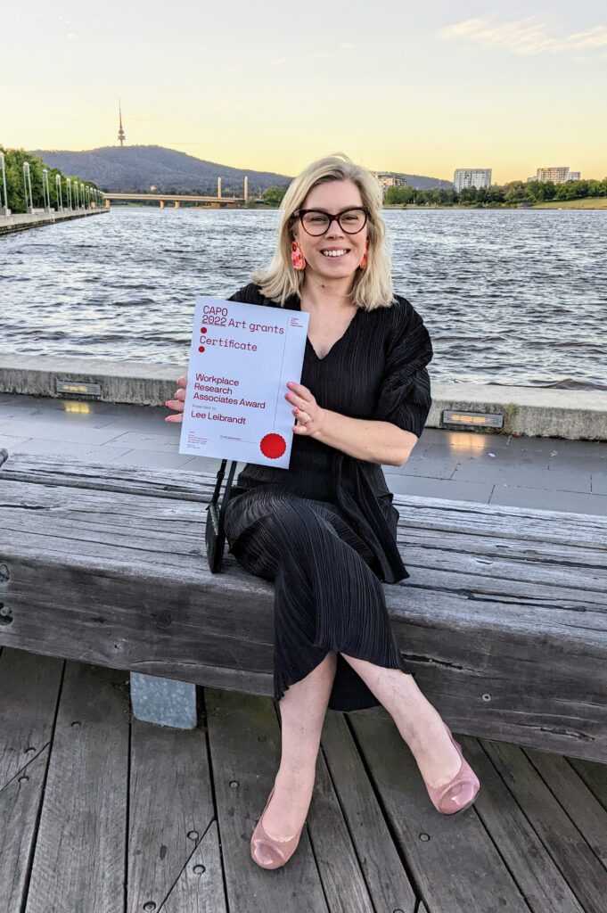 A smiling woman with blonde hair, glasses and big earrings, sits in front of a lake at sunset, proudly holding an award certificate.  