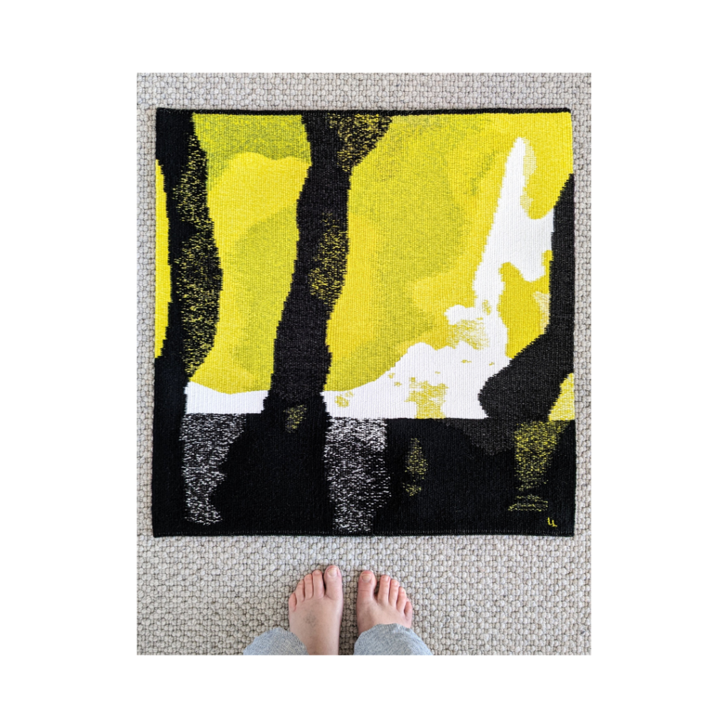 A large yellow, white and black woven tapestry lays on a grey carpet floor, with a woman's two feet below for scale.