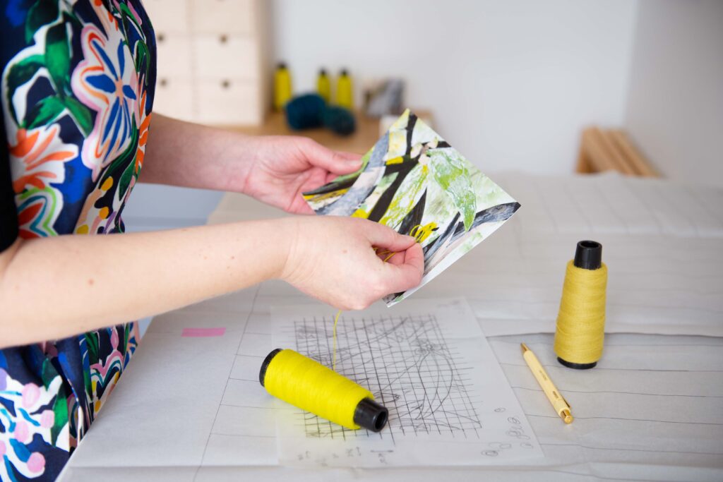 A white woman stands at a desk, holding a bright yellow thread of yarn up against a collage of eucalyptus leaves. Only her part of her torso and both arms are in the shot.