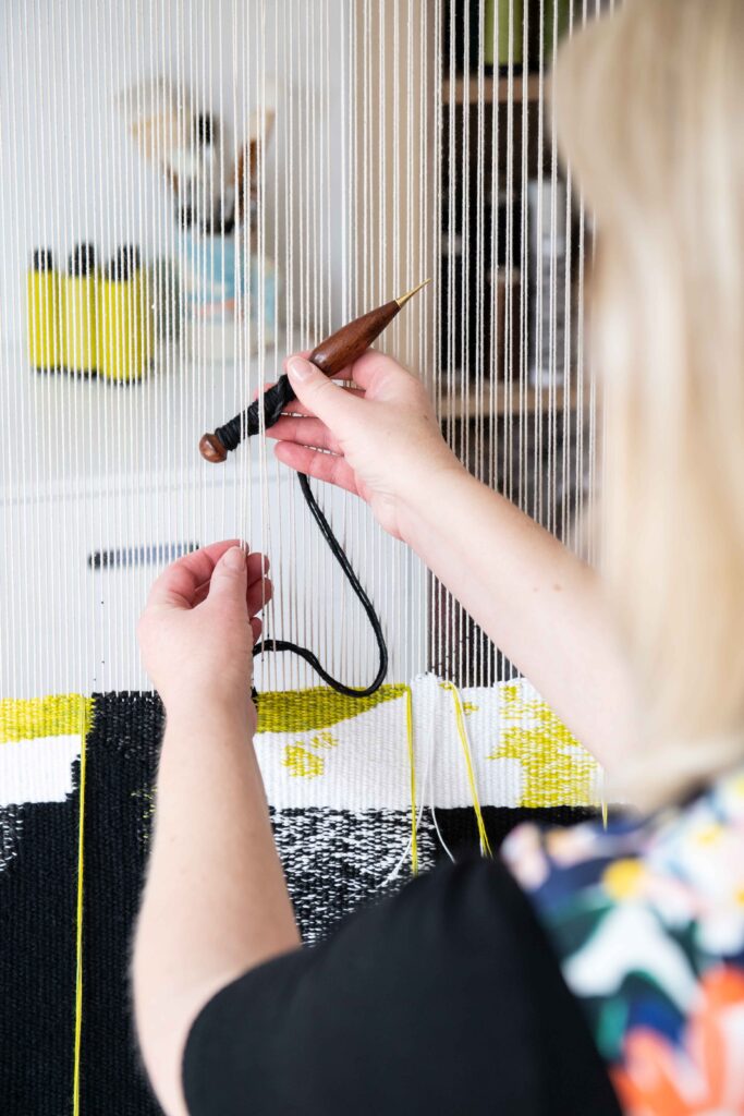 A blonde woman is weaving at a tapestry loom. She faces away from the camera, passing a tapestry bobbin through the warp of the tapestry. 