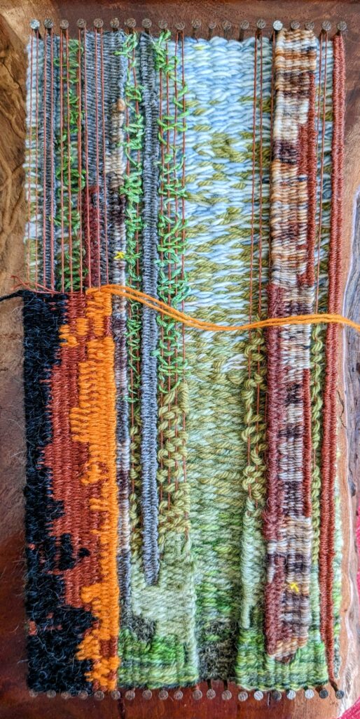 a woven tapestry of blues, greens, browns, grey, orange and black against a red gum timber block. Trees form within the tapestry.